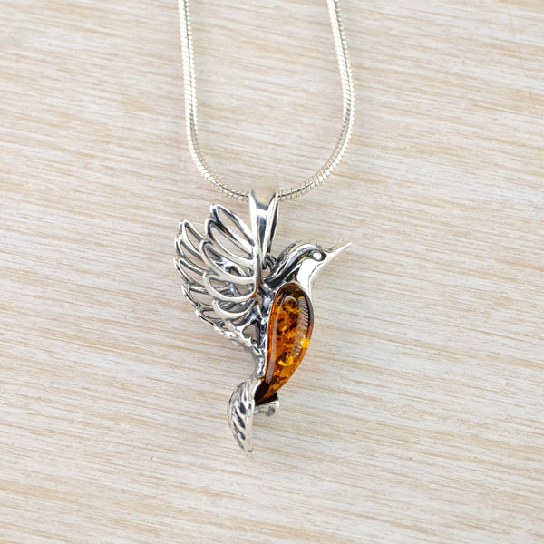A flying humming bird with silver wings with an open feather design and a silver tail. The body is deep orange amber with markings inside and the head is silver with the beak poised to seek nectar in a flower.