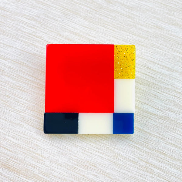 Bright square brooch. To the left is a bright orange square taking up most of the brooch. To the right is a gold  sparkly small rectangle, a white rectangle a blue square forms the corner. then to the left forming the bottom stripe is a white rectangle and a black rectangle . It forms a grid pattern.