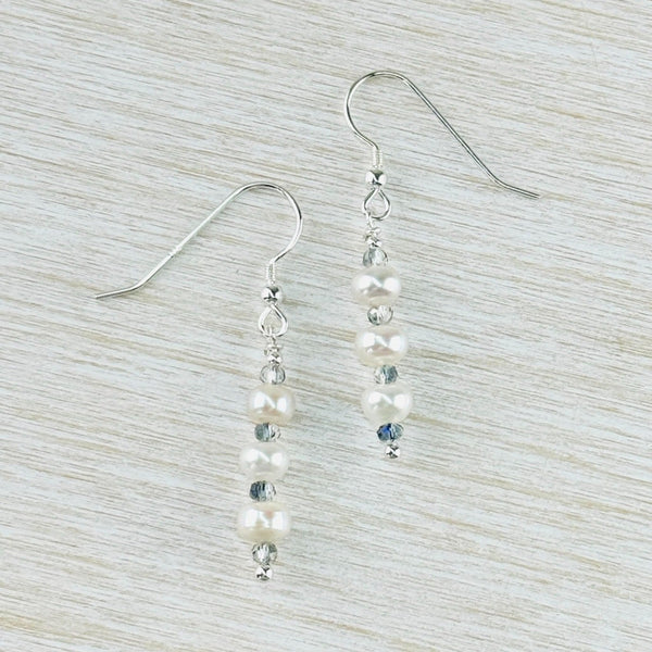 Long drop earrings with three round white pearl beads alternating with smaller pale grey  crystal beads. Hanging off a silver hook.