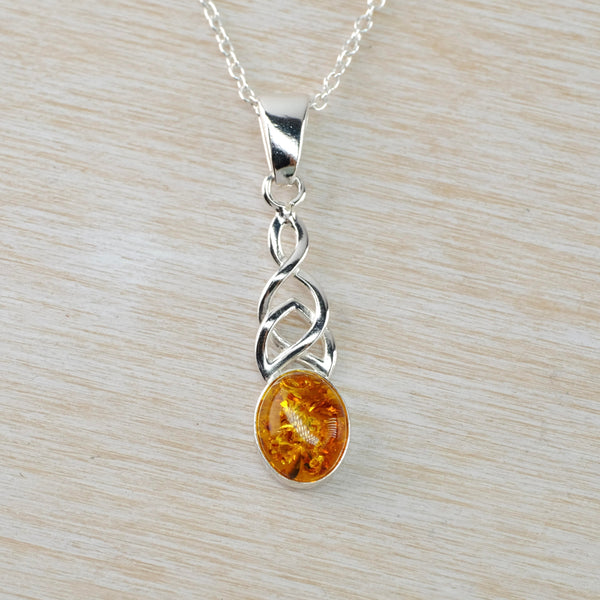 an oval piece of amber, framed in silver, sits below a simple silver celtic knot. The amber is dark orande with lots of internal markings some lighter, some darker. Hanging off a silver link chain.