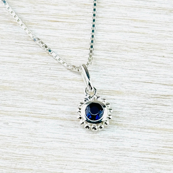 Small round pendant hanging from a  a square shaped shiny silver chain. The stone in the centre is clear dark navy blue. This is set in a silver circle surrounded by small silver balls.