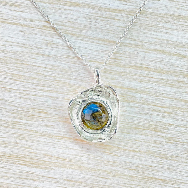 A pretty blue/ brown/ green round stone is set in a vaguely round, but organic shaped surround in textured silver. It hangs from a smooth bale on a dainty twisted chain.