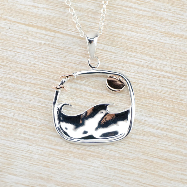 The shape of the silver pendant is a square with rounded corners. The bottom half is filled with ocean waves, top left are twogold coloured flying birds and top right is a gold coloured sun.