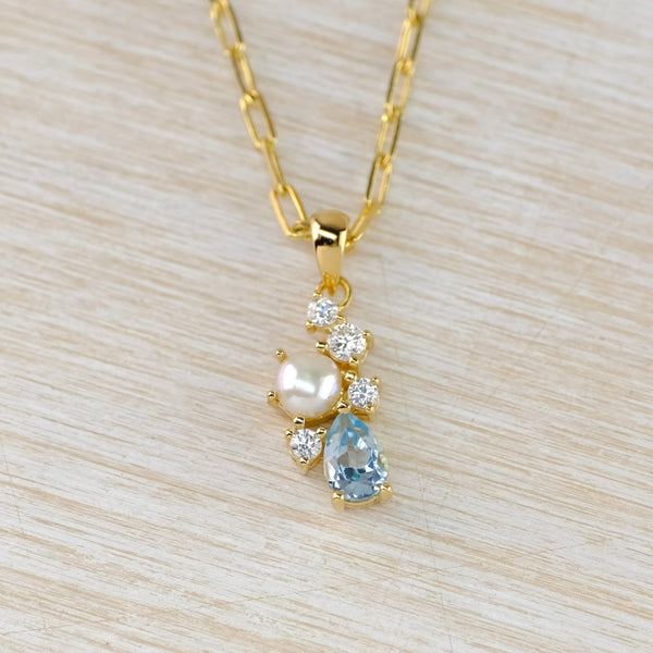 Gold plated pendant with a glossy round pearl  on one side a tear drop shaped sparkly blue stone on the other and four little clear sparkly stones winding between the two.