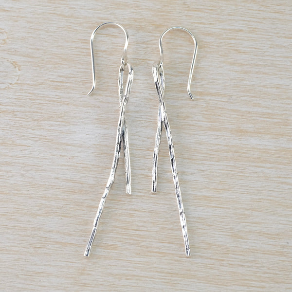 Earrings formed from long textured sticks that are folded back on themselves to form a loop for the hook to fit on. The piece at the back is approximately two thirds the length than the front. The sticks are shiny silver with a hmmered finish.