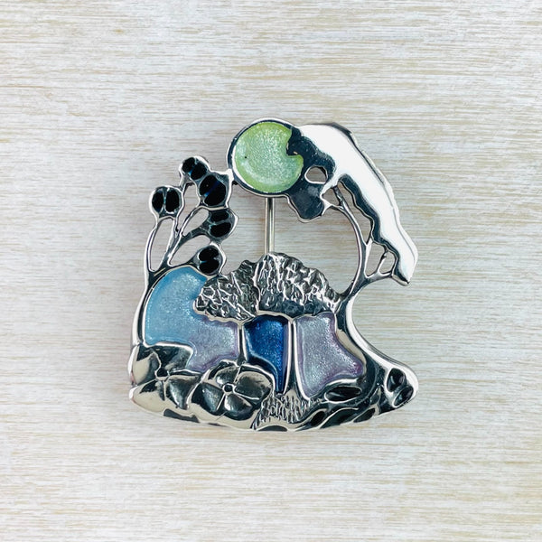Pretty woodland scene with shiny silver trees and flowers and infilled with pretty blue and green enamel.At the bottom of the brooch are two large 4 petalled flowers and some tree roots. Half way up are two small silver trees in full leaf, between these are three coloured sections. Two taller trees curve in towards the centre towards a pale green 'sun'. Very art nouveau style.
