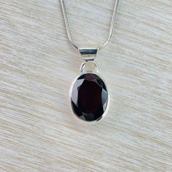 Deep red, faceted, oval shape garnet simply set in a silver surround with a circular ring hanging off a thick silver bale.