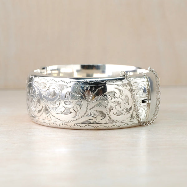 A rounded shiny silver bangle with a safety chain on one side. It has a delicate engraved design of swirls, dots and little leaves with a wavy design top and bottom.