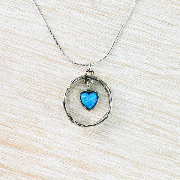 A vibrant light blue heart shaped opal  is set in silver and hangs from a silver ring within a larger silver circle of textured and darker silver. Hanging from a shiny silver chain.