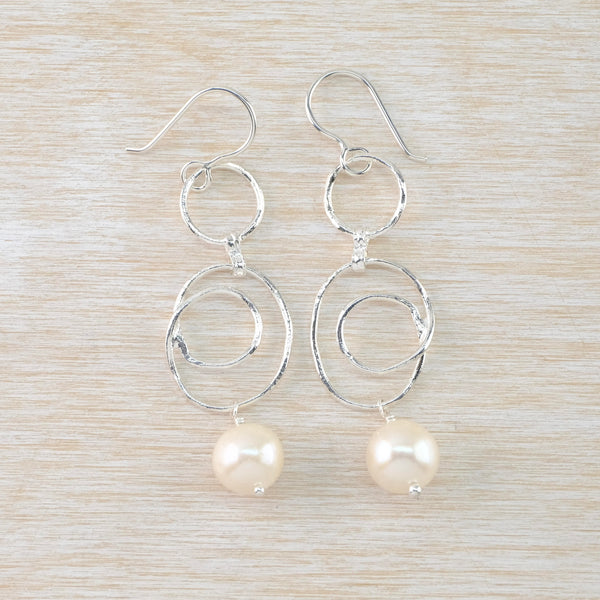 Long textured silver earrings hanging from a silver hook. A round creamy pearl hangs from an open twisted circle which itself hangs off a smaller open circle.