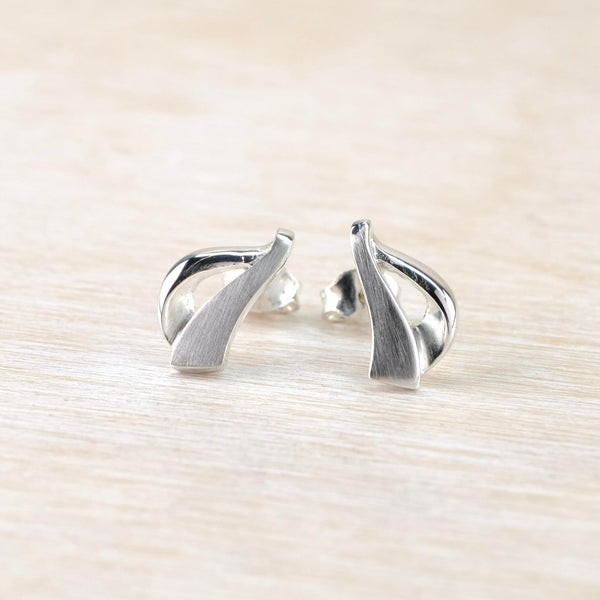 Vaguley in the shape of a harp. One side of the earring is a flat satin silver shape, the other side is a curved shiny silver which is attached to the satin silver at top and bottom.