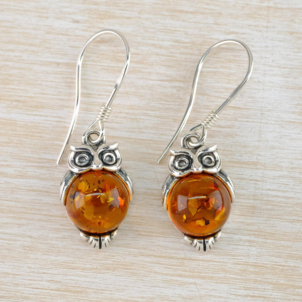 Owls as earrings. They have large round amber tummies in a deep orange colour with markings in the amber. Wide silver eyes are on he top and short silver wings around the top half. They are finished with silver feet on the bottom.