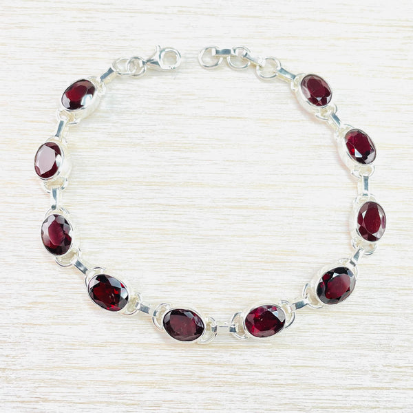 Ten deep red sparkly oval stones, set in silver. Each stone link has semi circle pieces of silver attached at each side. Between each link is a short rounded rectangular link. It has a lobster catch at the end and a plain piece of chain at the other side.