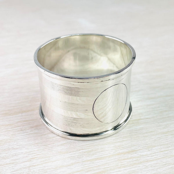 Round napkin ring with shiny rims top and bottom. The decoration is three sets of horizontal stripes., ten in each group . A round plain cartouche is  empty at the front.