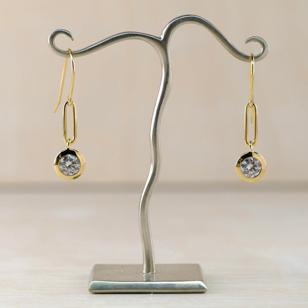 A circular sparkly clear stone hangs at the end of these earrings. It is set in a gold frame with a long slim rectangle with rounded corners above it All hanging off a gold hook.