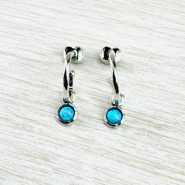 High polished silver in a three quarter hoop design with a silver ball at one end and a post and butterfly fitting at the other. Suspended from this is  a round bright blue opal stone set in a darker grey silver frame.
