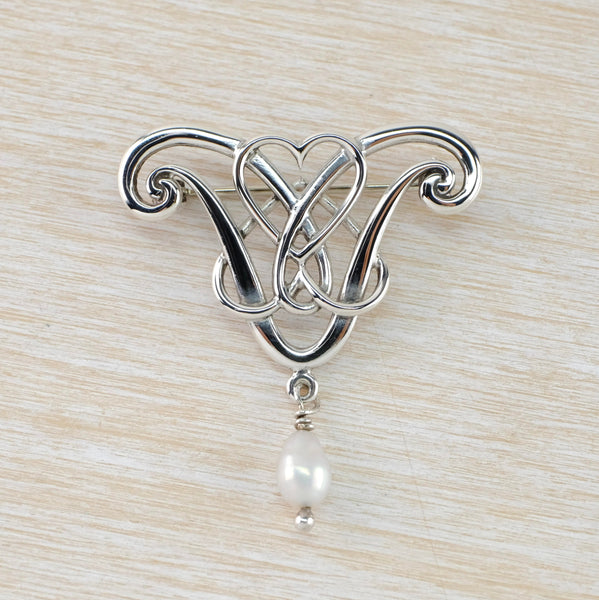 Ornate silver brooch with a real celtic look. In the centre are two intermoven silver hearts, surronded by an ornate vaguely heart shape. I white egg shaped pearl dangles from the bottom.