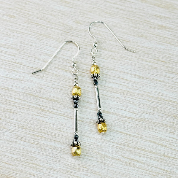 Long earrings hanging off a silver hook. Top and bottom are gold coloured textured round beads, then dark silver little balls around the stem, then small dark grey beads - the design mirrors itself. Between this is a shiny silver rod to give the length.