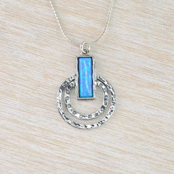 A bright blue rectangular opal stone is set within a texture silver frame. Two hammered silver circles, one larger than the other are attached to the rectangle - about half way down. These extend past the bottom of the rectangle.