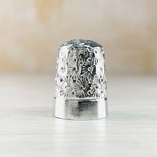 The top two thirds of the shiny thimble are decorated witha repeated flower design, a bit like a chrysanthemum. The bottom third is plain high pokished silver.