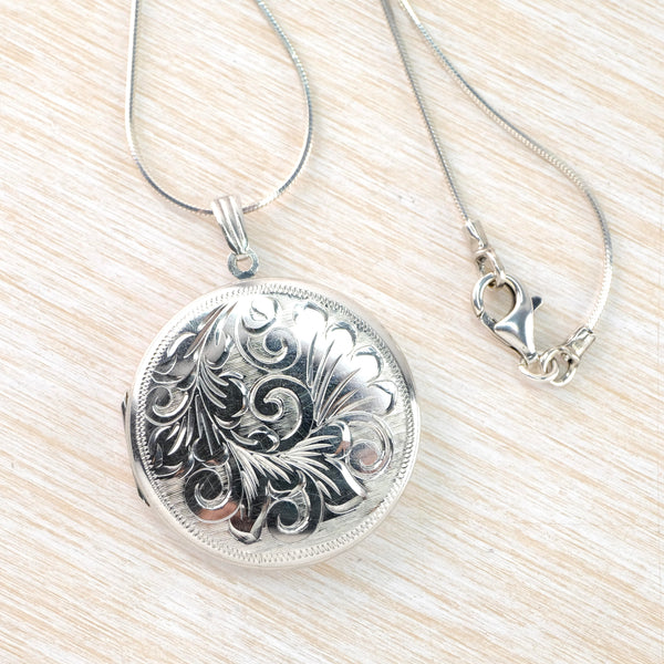 A round shiny silver locket. The front is decorated with swirls and fleur de lys details, framed by a fine wiggly design and then a plain silver edge.