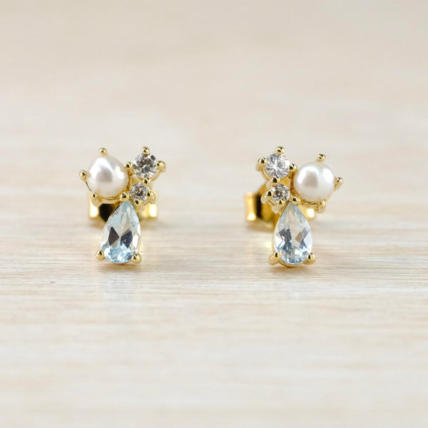 Gold coloured earrings with a sparkly blue tear drop stone at the bottom, a round glossy pearl to one side and two smaller sparkly clear stones to the other.