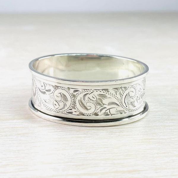 A bright silver oval shaped ring. Plain silver rims with an additional plain silver band just inside the rim. The rest of the napkin ring is engraved with swirls formed of dots and shadings made of short stripes.