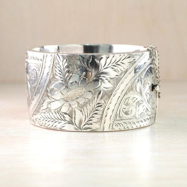 Shiny silver bangle with a safety chain on one side. It is deep in size with a central flower and leaf design surrounded by swirls, dots and little leaves.