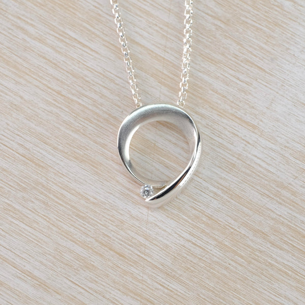 An open circle design with a little 'tail' at the bottom, pointing to the left. There is a little sparkly Cz stone nestling between the circle and the tail. The circle has a satin finish with  high polished silver edges. Hanging of a delicate silver link chain.