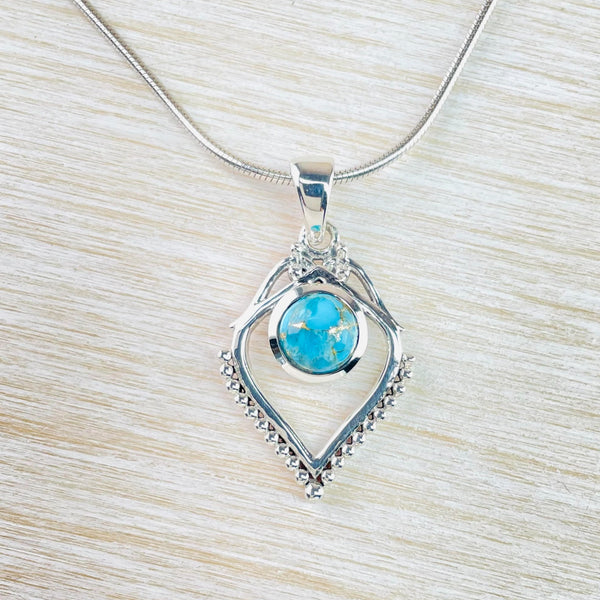 Overall diamond shape hanging from a silver hook. At the top of the diamond the silver is crossed over to join the hook. Below this round bright blue turquoise stone, set in silver, are set centrally but a little above the half way point length wise. The bottom half of rhe diamond has tiny silver balls along the outside. Hanging from a silver chain.