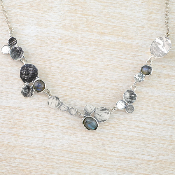A half necklace formed of randomly placed textured silver disc, in groups of two or threes.  There are differing sizes of discs with three different sized dark grey/green labradorite stones randomly included. The back of the necklace is a fine silver link chain.