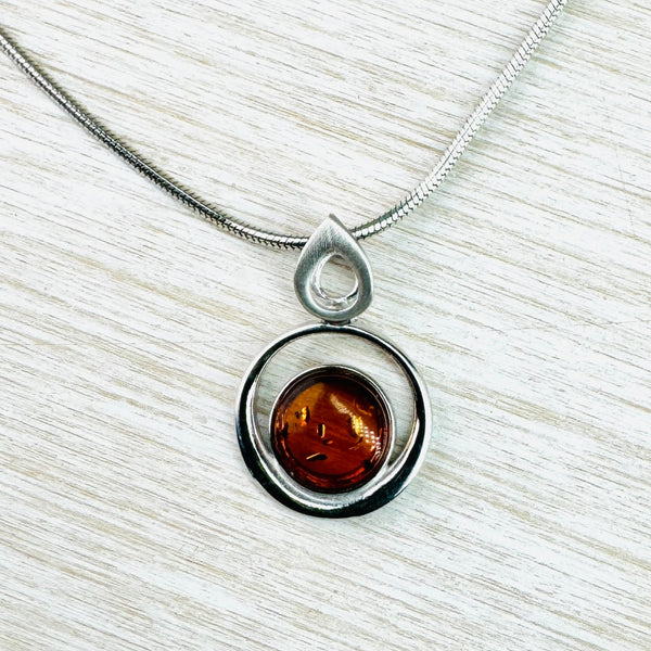 A deep orange amber stone, with a few delicate markings, sits at the bottom of an open silver circle,leaving space to see through. The circle hangs of a satin silver open tear drop shape which the chain threads through.