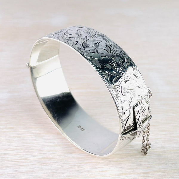 Bright silver bangle, one side decorated, the other plain and high polished. The decorated side has a dotted edging and is engraved with fleur de lys style patterning. The catch is decorated with four crosses.