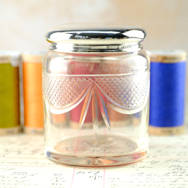 A jar shaped glass pot, topped by a shiny silver lid. The glass is decorated with deco style swags and bows design in the top half, plain at the bottom.