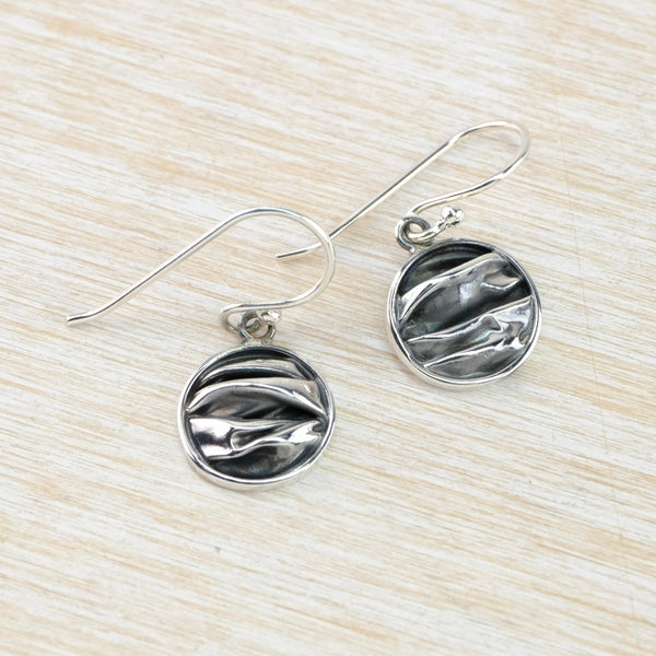 Round earrings hanging off a silver hook. They have a high polished frame and the inside has a dark grey background with bright silver in the raised folded stripes.