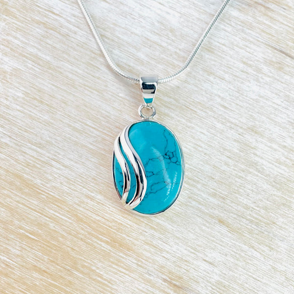 Blue turquoise oval stone framed in silver with two wavy shiny silver stripes  on top of the stone offset to the left hand side of the stone.