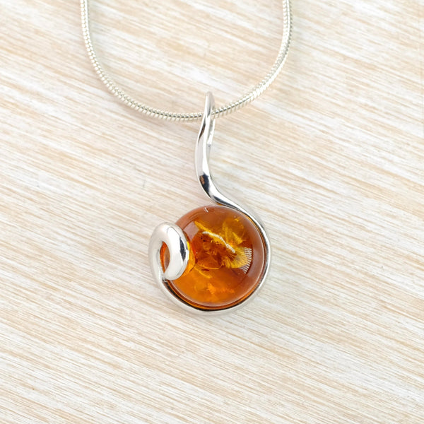 A lovely orange amber stone with little seed pods enclosed.The amber is round and is enclosed in silver three quarters of the way round. The silver then gently twirls across the front of the side of the amber. The silver looksmlike an upside down question mark as it connects to the chain.
