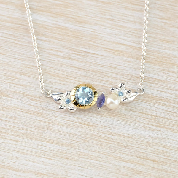 The necklace has a pendant attached vertically to a chain. A silver flower is at each end, with a tiny blue stone in the middle. Left of centre  is a larger faceted blue stone set in gold, a purple diamond shaped stone and then a glossy round white pearl.