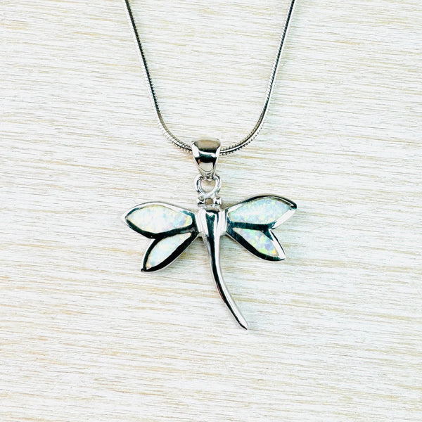 Silver dragonfly has a silver body, curving slightly towards the bottom. Two wings on each side have silver sedges filled with sparlky stones which show colours of cream, green and pink. It hangs of a plain silver bale on a chain.