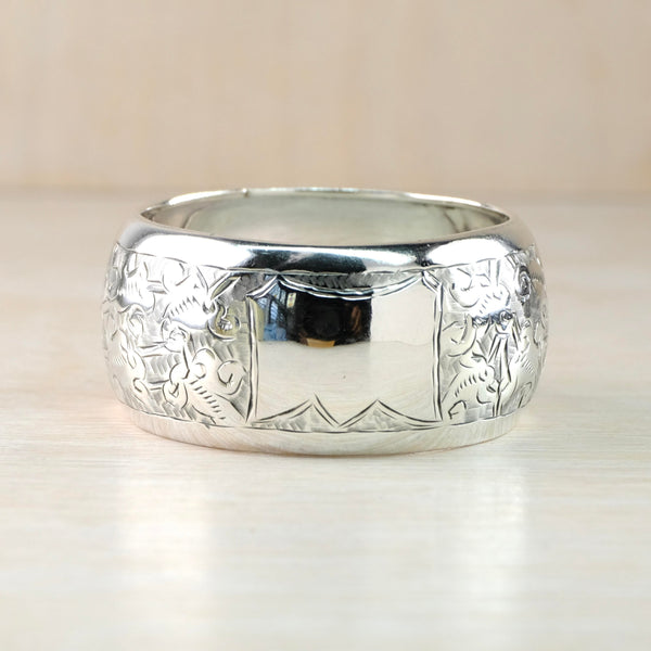 Round convex shaped napkin ring in very shiny silver. In the centre front is a plain silver rectangle surrounded by delicate engraving of flowers and swirls.
