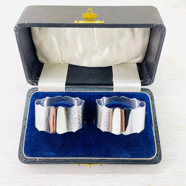 A black leather box with a blue velvet lining. Inside are two identical napkin rings with wavy edges. An engine turned fine wavy line decoration goes all the way around maybe 20 lines deep. At the front is a shiny silver blank space (for engraving) 