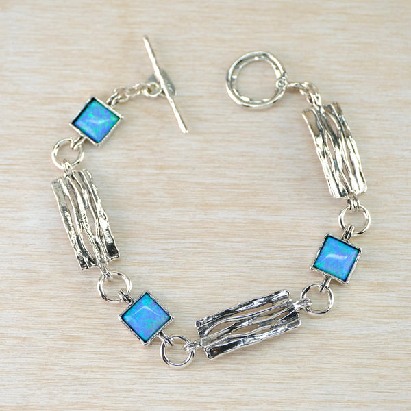 Three pretty bright blue square opal stones, framed in silver, alternate with three rectangular silver links - about three times as long as the opals. The silver links are decorated with irregular ridges along their length. The links are connected by rpond silver jump rings and it is finished with a shiny silver T bar and circle.