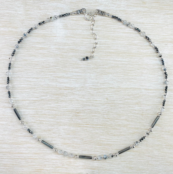 A pattern of grey, black and pale blue beads repeats around the necklace. Small round opaque shiny grey beads, long tubular grey beads, round pale blue beads and pale grey beads with black flecks. Round silver beads are dotted within.