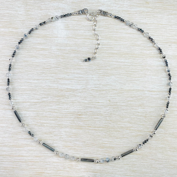 A pattern of grey, black and pale blue beads repeats around the necklace. Small round opaque shiny grey beads, long tubular grey beads, round pale blue beads and pale grey beads with black flecks. Round silver beads are dotted within.