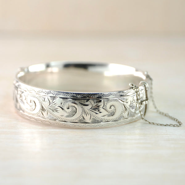 Pretty shiny bangle withleaf and fleurs de lys style ehgravings around the front. The top and bottom have a different design - looks a little like a zip pattern within a stripe. Safety chain to the right.
