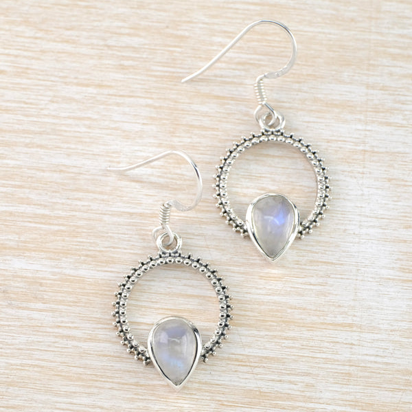 Drop earrings with a silver hook. The drop is formed of a decorated silver circle with little silver blobs inside and out. At the bottom of the circle a tear drop moonstone in a silver surround, is attached, half above and half below the circle .