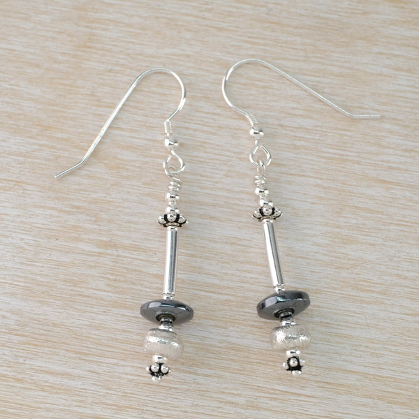 Long earings with a disc shaped shiny grey hematite bead above a textured round silver bead at the bottom. Above this is  a long shiny silver tube and amore small silver beads. Hanging off a silver hook.