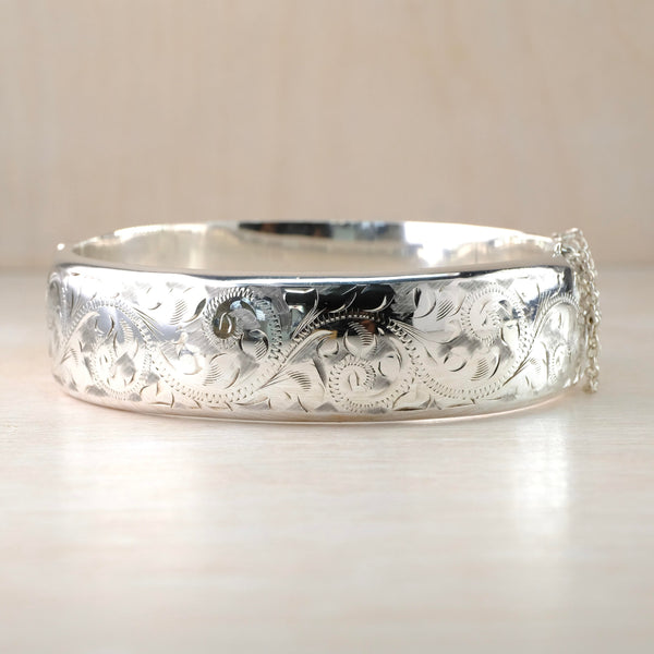 Shiny silver bangle with a safety chain on one side. It has a delicate engraved design of swirls, dots and little leaves.