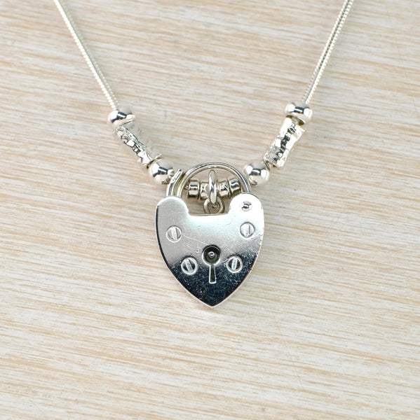 A shiny silver heart shaped padloc pendant with four screwheads on the front. Threaded onto a chain with mixed silver beads threaded along the first inch on either side nearest to the pendant.