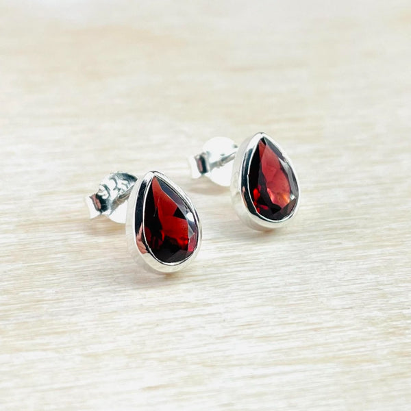 A pair of teardrop-shaped stud earrings with deep red faceted garnet stones set in silver, displayed on a white wooden surface.